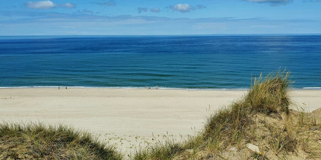 Blick von einer Düne hinunter auf den breiten Sandstrand und das azurblaue Meer.